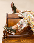 "Woman reclining in a chair wearing 'Liz' loafers in Army Leather with Inversa Python accents, paired with vibrant, patterned trousers for a bold fashion statement."