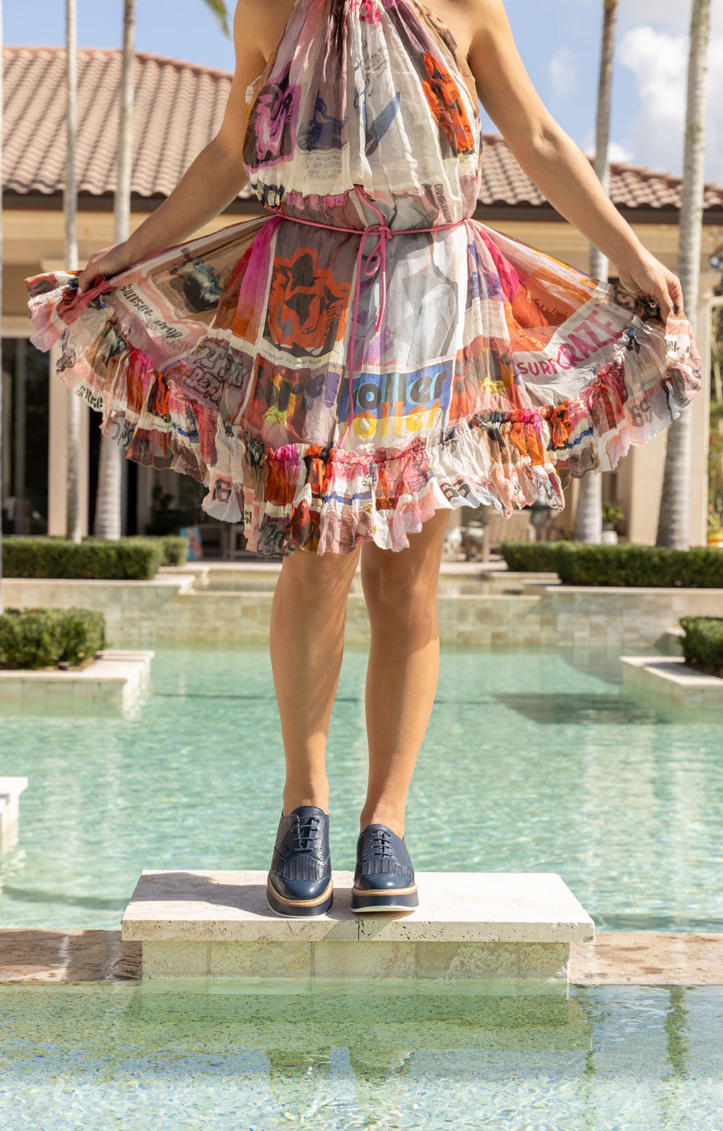 Woman standing on a stone ledge by a pool wearing &#39;Liz&#39; Oxford shoes in Nuit Leather, paired with a colorful, airy dress, showcasing a blend of casual elegance.