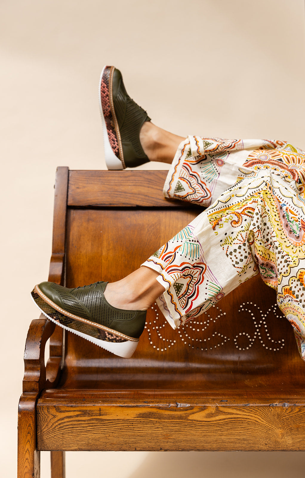 "Woman reclining in a chair wearing 'Liz' loafers in Army Leather with Inversa Python accents, paired with vibrant, patterned trousers for a bold fashion statement."