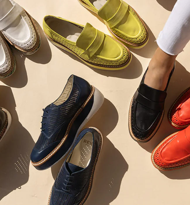 Assortment of Rex Shoes' luxury loafers for women in various colors, including classic white, vibrant red, and chic metallics, displayed around a stylish woman dressing up on a sunlit background. - Cropped to frame just the woman's foot with five pairs of shoes around her.